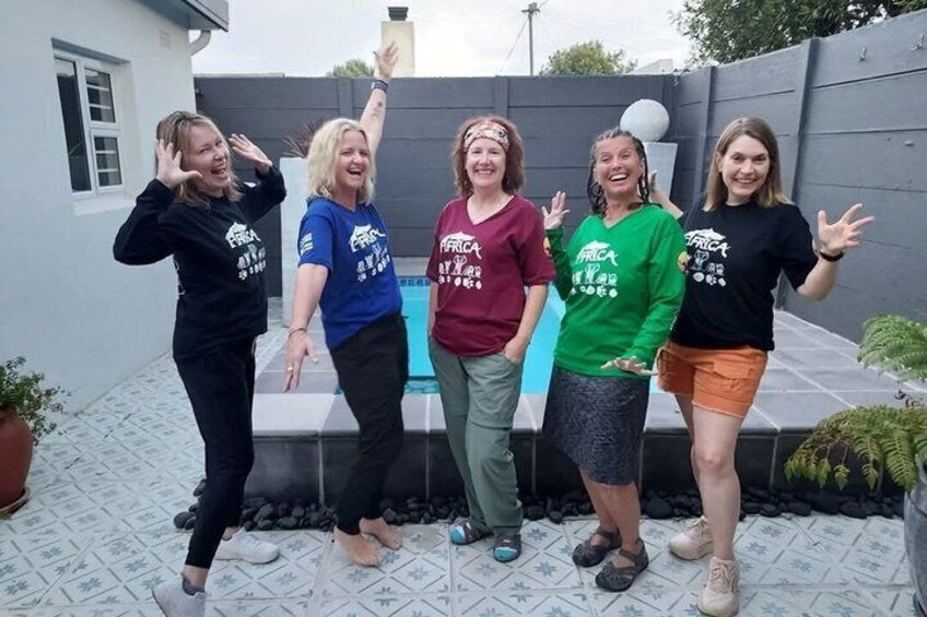 Vier Personen stehen an einem Pool mit gefliestem Patio, gekleidet in Freizeitkleidung und T-Shirts mit dem Aufdruck 'FRICA'. 4