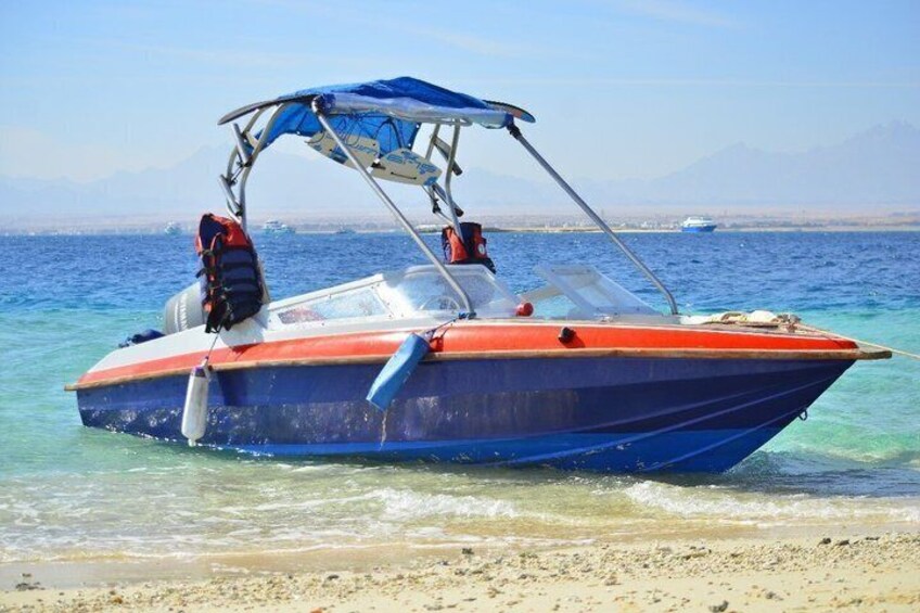 A small boat with a blue and red hull, a canopy, and life jackets, anchored near a sandy beach with clear water and distant mountains. 1