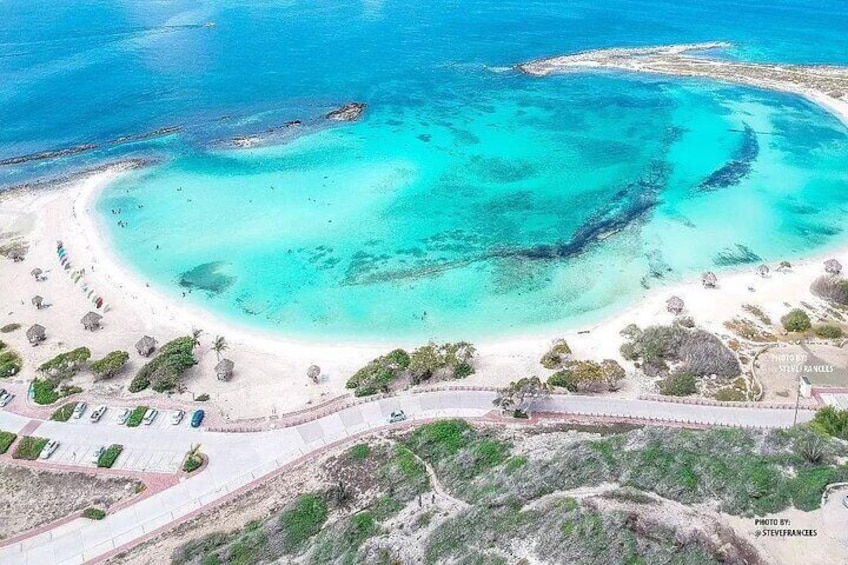 A coastal landscape with a sandy beach, clear turquoise waters, and a small island with rocky outcrops. 1