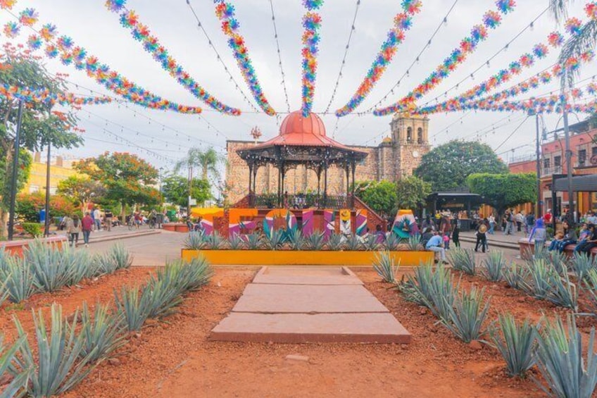 Una plaza con un quiosco, guirnaldas de flores coloridas y un edificio histórico al fondo. 3