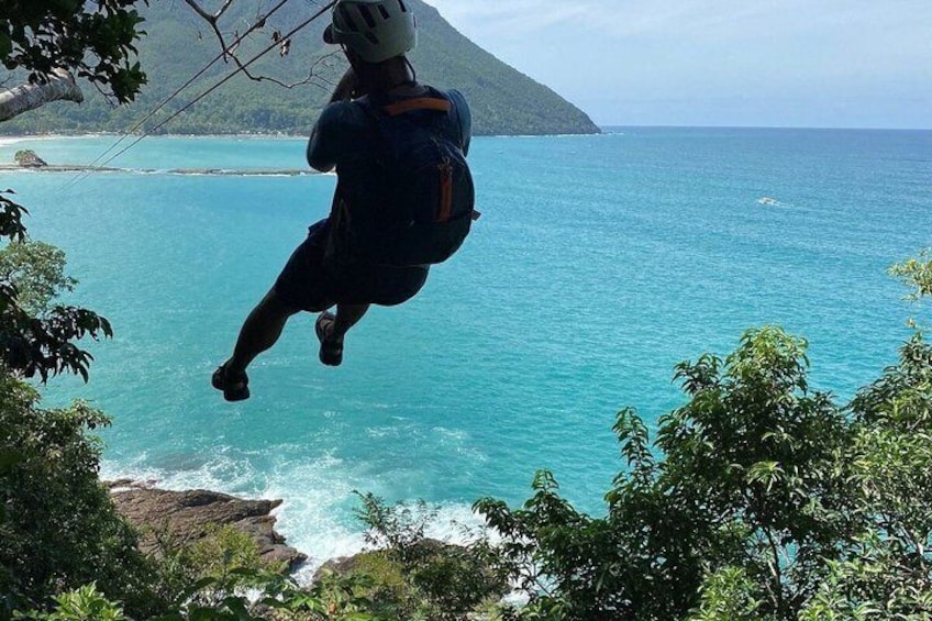 En person er i gang med at bungee hoppe med en rygsæk, med udsigt over et turkis hav og et bjergrigt landskab. 4