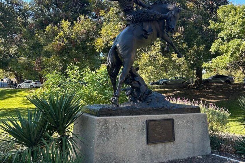 A bronze equestrian statue on a pedestal in a park with trees and parked cars in the background. 3