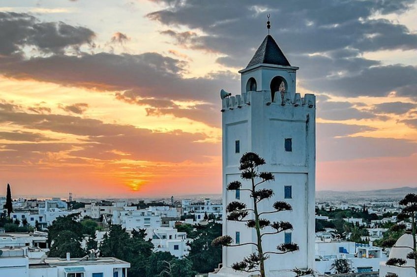 A white tower with a pointed roof, surrounded by buildings and trees, during sunset. 2