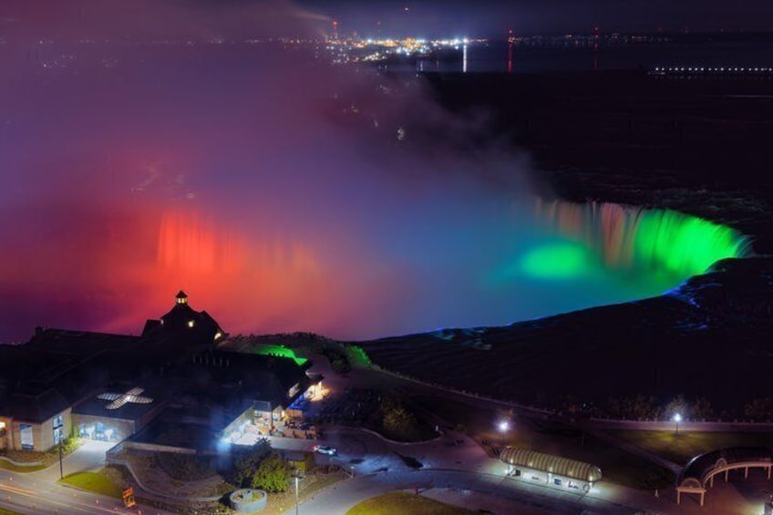 A nighttime view of a waterfall with colorful lights, a dome-shaped building, and a lit parking area. 3