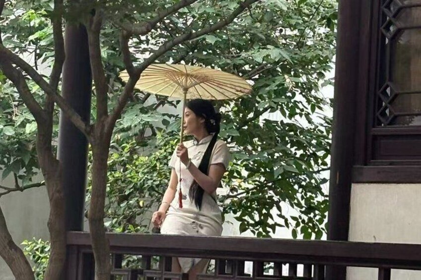 Une personne se tient sur un balcon, un parapluie à la main, avec des arbres et un bâtiment en arrière-plan. 1