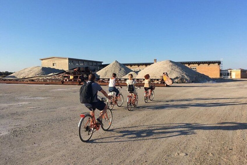 People riding bicycles on a gravel surface with industrial buildings and piles of sand in the background. 3