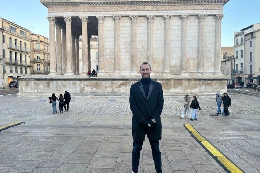 A man standing in front of a classical building with columns, with people walking in the background. 4