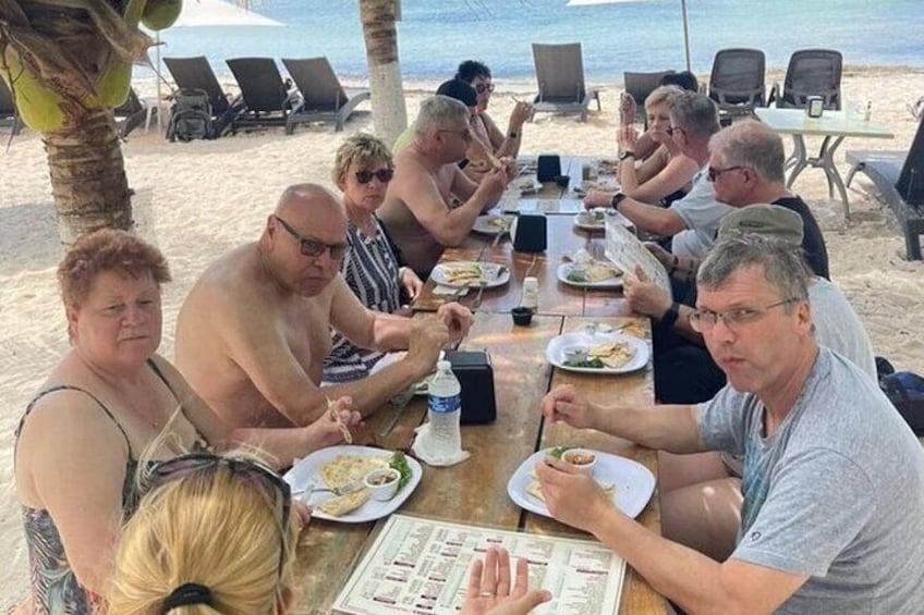 En grupp människor sitter och äter vid ett träbord på en strand, med palmer och strandstolar i bakgrunden. 1