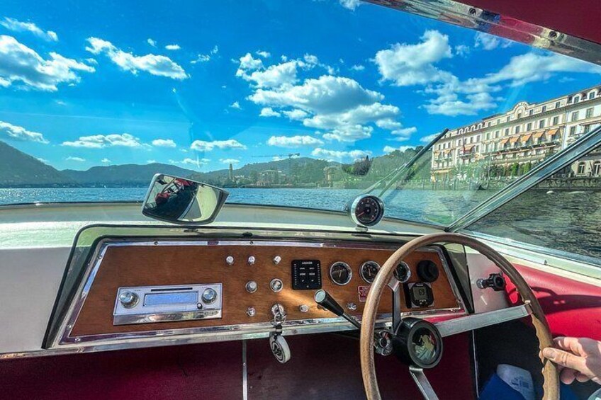 Interni di una barca con timone, cruscotto e vista su un lago con edifici sullo sfondo. 5