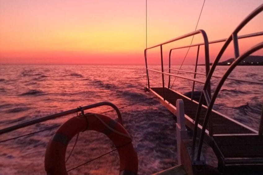 A boat deck with a lifebuoy and railing, overlooking a sunset over the ocean. 2