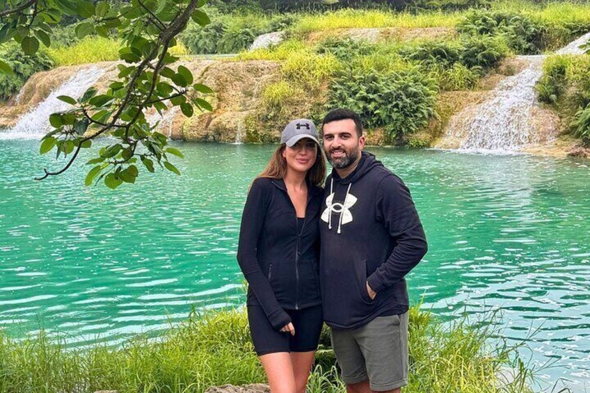 Two people standing by a turquoise lake with a waterfall in the background. 1