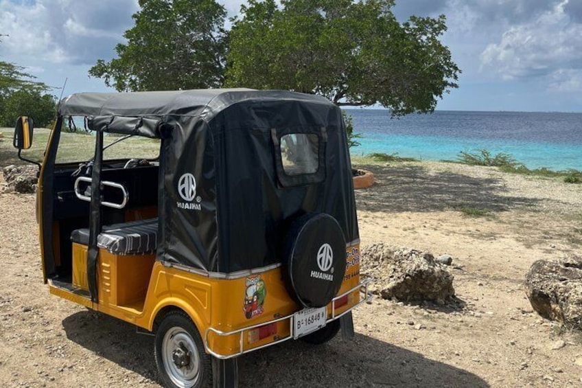 Un tuk-tuk amarillo y negro estacionado en una playa arenosa, con vista clara al océano y árboles al fondo. 5