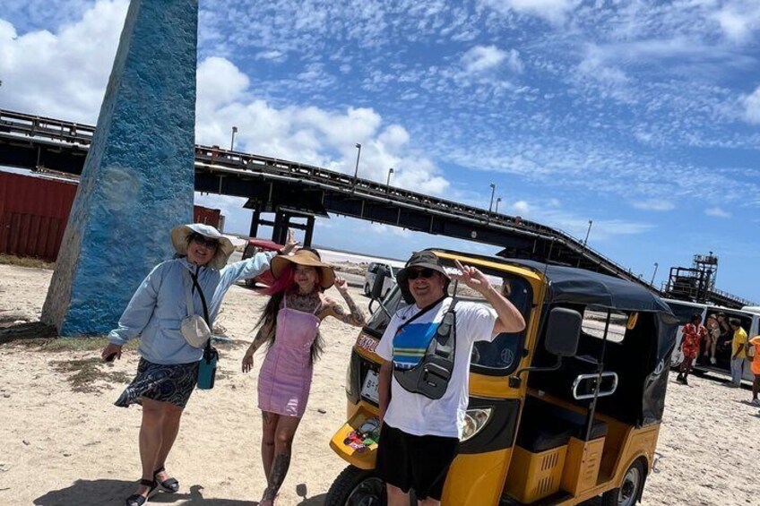 Tres personas de pie junto a un auto-rickshaw en una playa arenosa, con un puente al fondo. 4