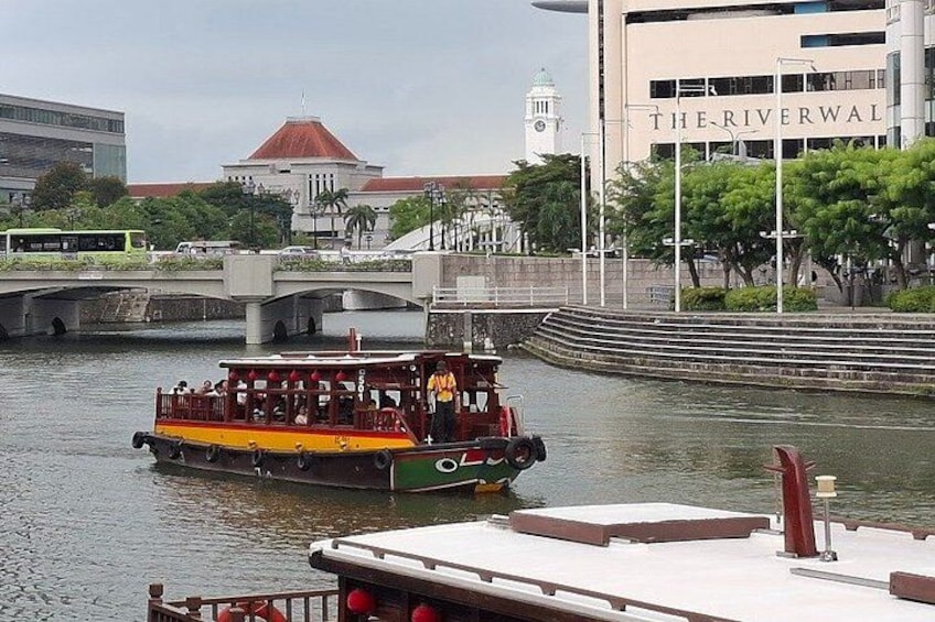 Ett flodbåt med passagerare, en bro och en byggnad med texten ”THE RIVERWAL” på, syns i bakgrunden. 5