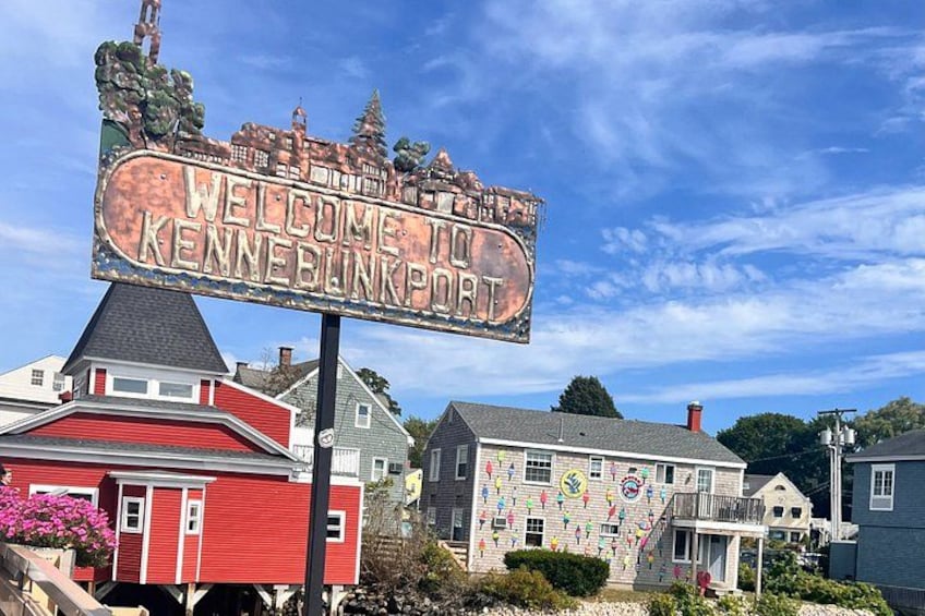 Un cartello di benvenuto per KenneBlinkport, circondato da case con decorazioni colorate. 1