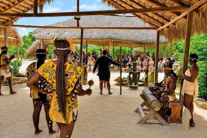 A cultural event with people in traditional attire, a thatched-roof structure, and a sandy ground. 1