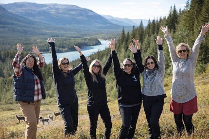 A group of people standing on a grassy hill with their hands raised, overlooking a lake and mountains in the distance. 1