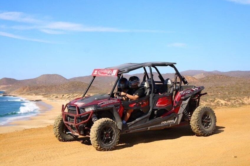 A Polaris off-road vehicle on a sandy beach with ocean and hills in the background. 1
