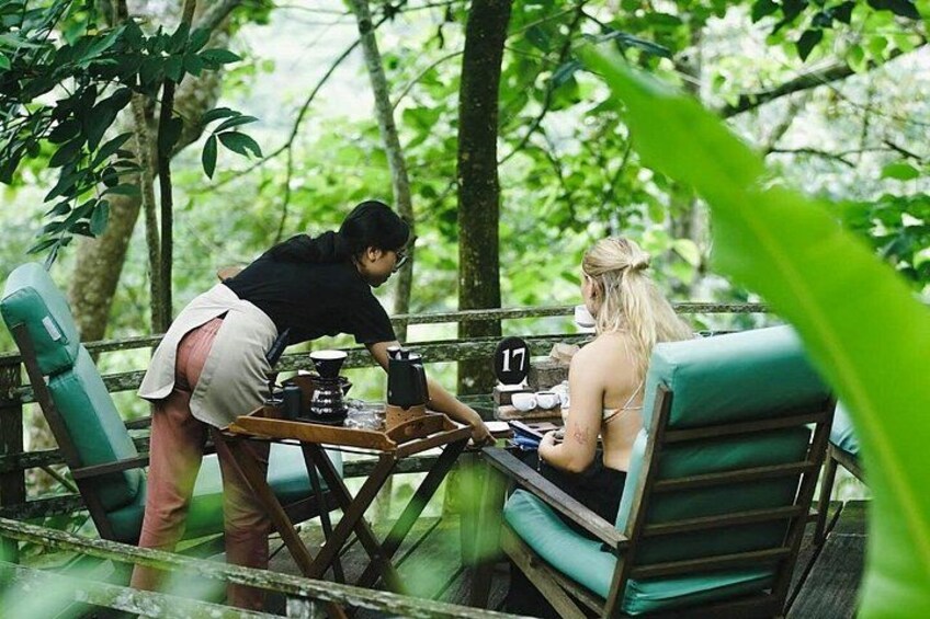 Dos personas sentadas en una terraza de madera en un área boscosa, con una mesa entre ellas. 2