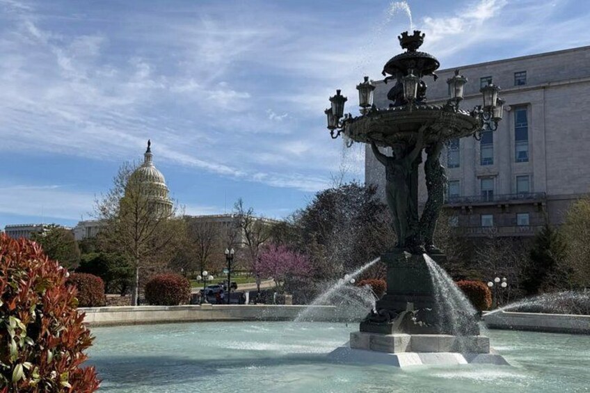 A fountain with water jets, a large building resembling a capitol in the background, and a clear blue sky. 2