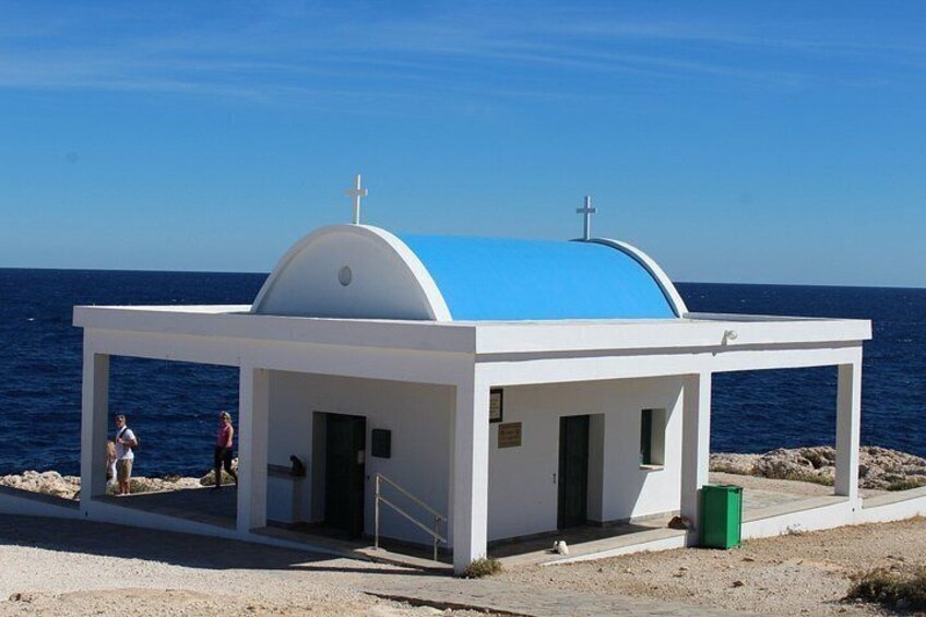 A small white building with a blue roof and a cross on top, located near a beach with the ocean in the background. 2