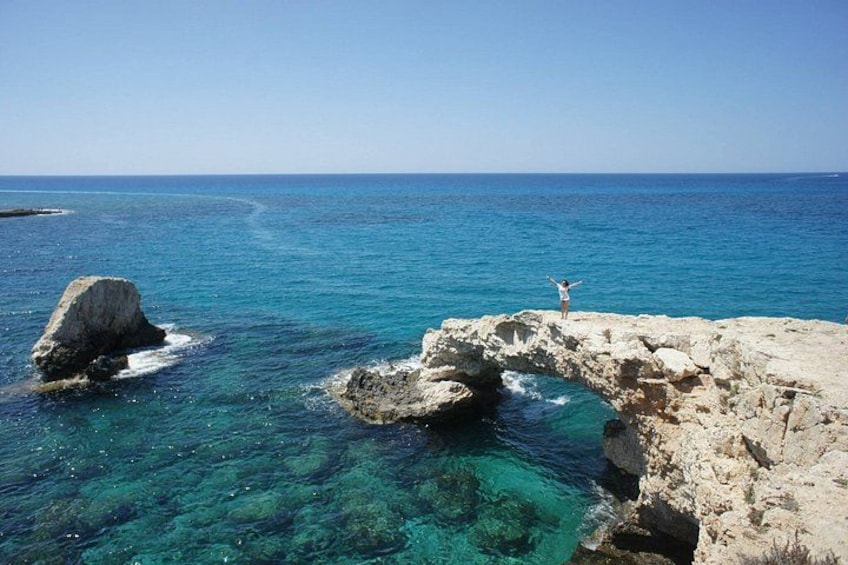 A rocky coastline with clear blue waters, a person standing on a rock, and a large rock formation in the sea. 4