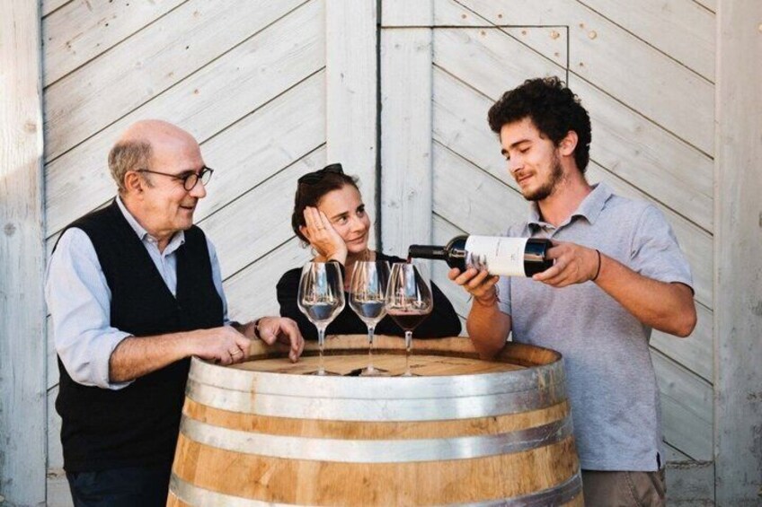 Three people at a wooden barrel with wine glasses, one person pouring wine. 1