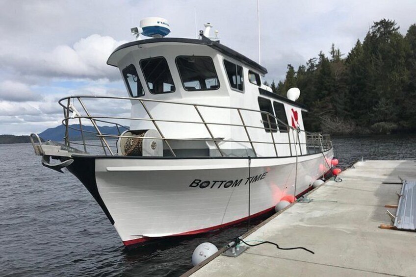 A boat named 'BOTTOM TIME' docked at a pier with a forested shoreline in the background. 3