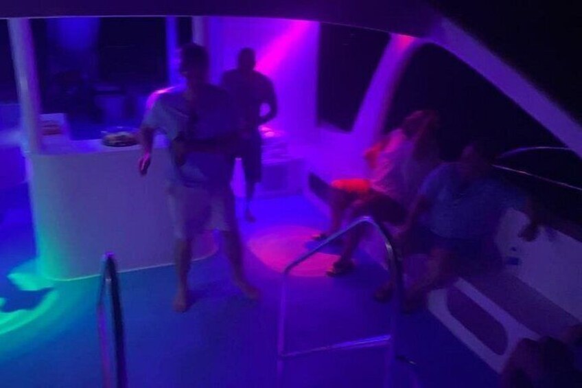 A dimly lit room with people standing and sitting, a bar area, and a staircase. 2