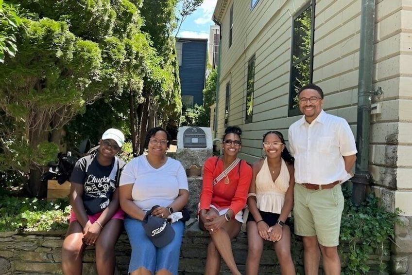A group of people sitting and standing on steps in front of a building with greenery around. 4