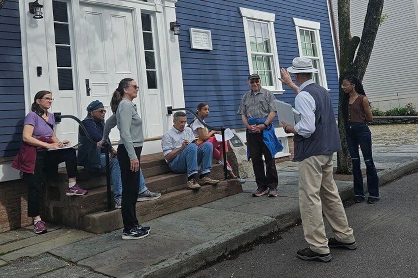 A group of people standing and sitting on a sidewalk in front of a blue house with white trim. 1