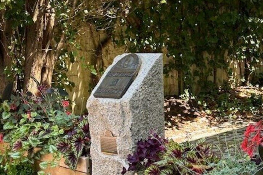 Monumento de piedra con una inscripción, rodeado de vegetación y un muro de piedra. 5