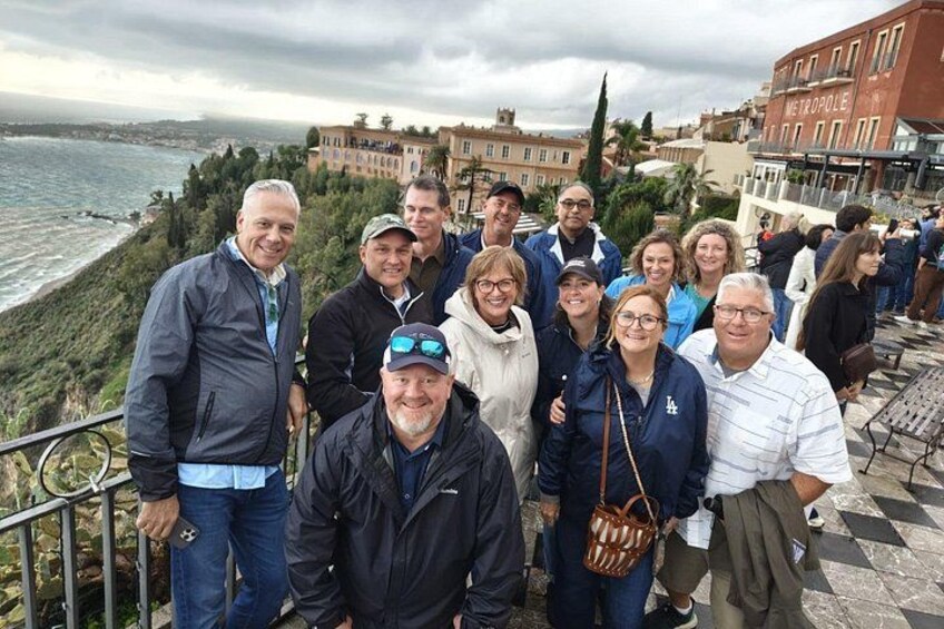Eine Gruppe von Menschen steht auf einer Terrasse mit Blick auf eine Küstenlandschaft, im Hintergrund sind Gebäude zu sehen. 4