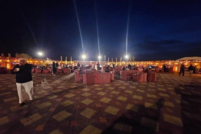 A nighttime outdoor event with illuminated tables and chairs, a checkered floor, and a crowd of people. 3