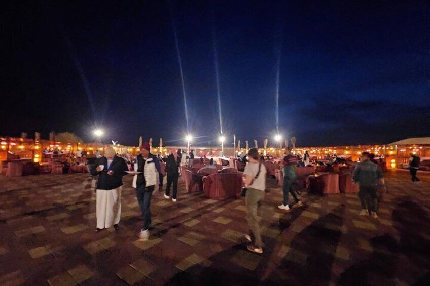 A nighttime outdoor event with people gathered around tables, illuminated by overhead lights. 4