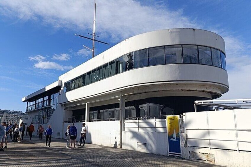 A modern building with a curved facade and large windows, located near a waterfront with people walking around. 2