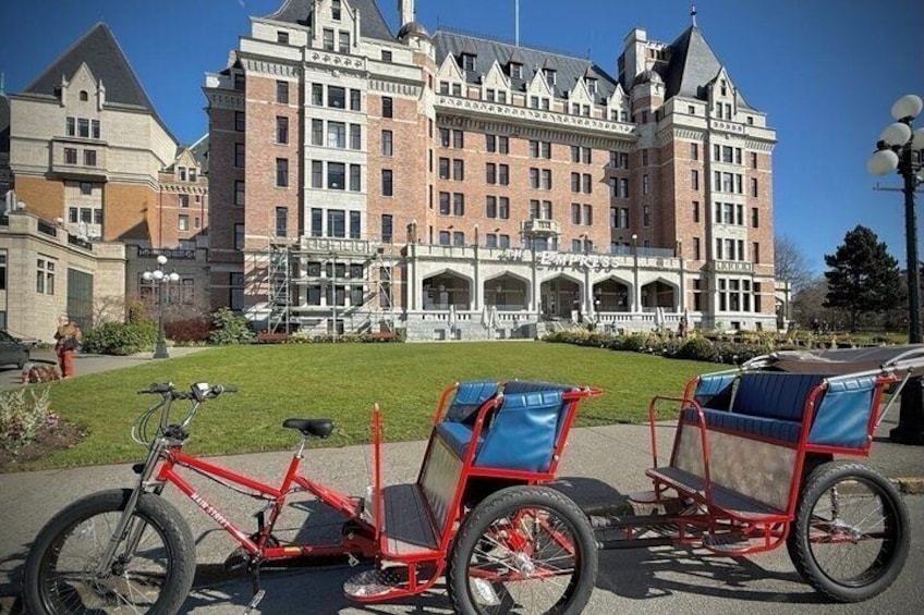 Una bicicleta roja con un remolque de carga estacionada frente a un edificio grande e histórico con fachada de ladrillo y múltiples ventanas. 1
