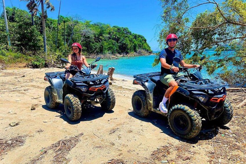 Two people on ATVs on a sandy beach with a clear blue sea and green foliage in the background. 4