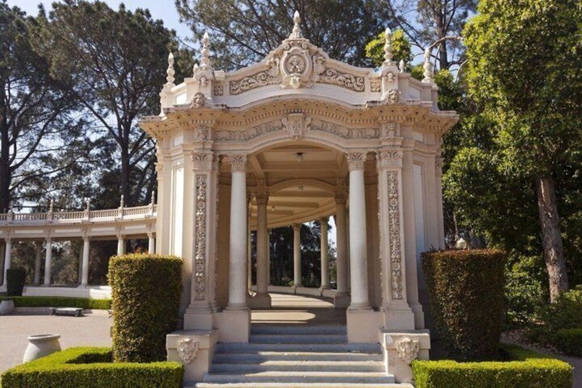Un padiglione classico con colonne elaborate e un balaustra, circondato da siepi curate e alberi. 2