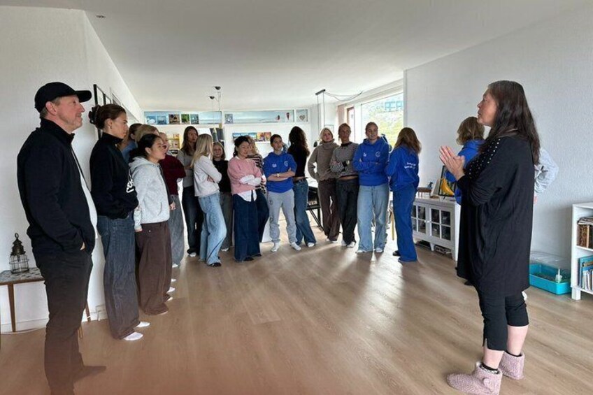 A group of people in a room with wooden floors and white walls, some standing and others seated. 5