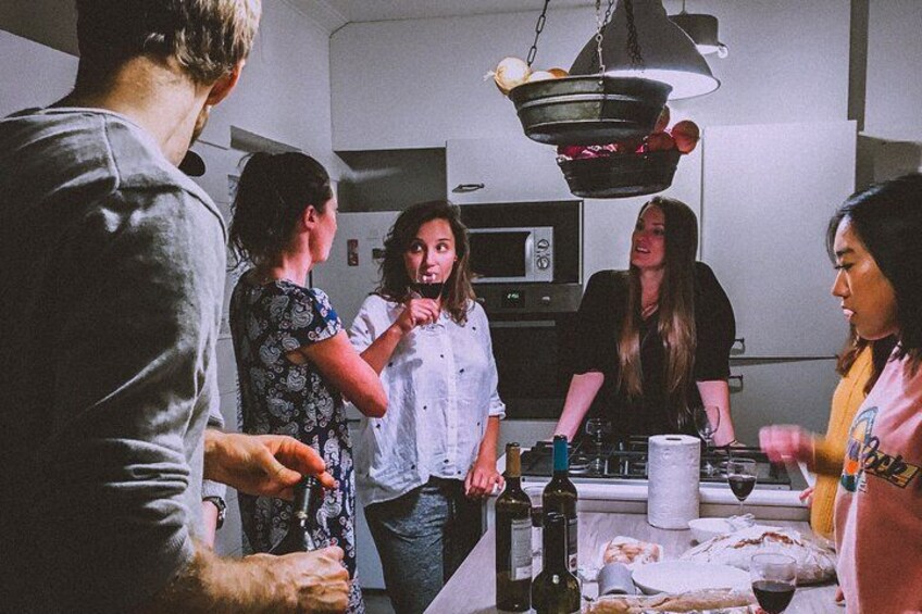 A group of people in a kitchen, some standing and some sitting, with a microwave and a hanging pot rack visible. 2