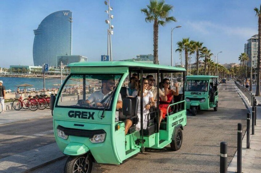 A green tour bus with passengers, palm trees, a beach, and a modern building in the background. 2