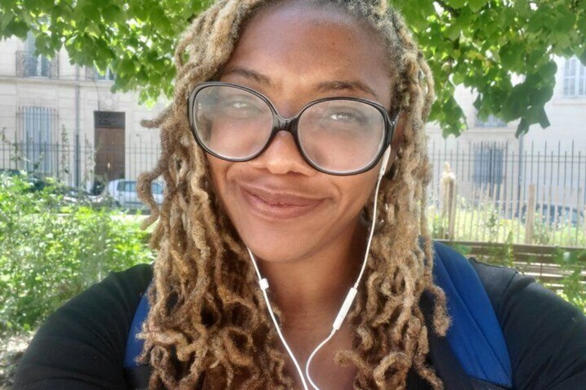 A person with curly hair wearing glasses and a blue top, standing outdoors with trees and a building in the background. 2