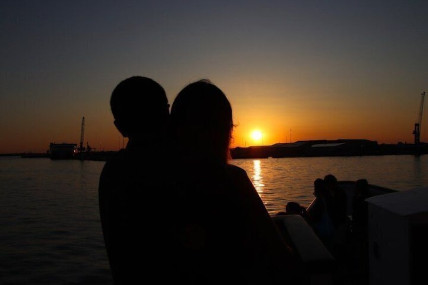 Un couple se détache sur le fond d’un coucher de soleil, avec une étendue d’eau et des bâtiments industriels en arrière-plan. 1