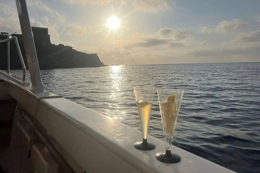 Una barca con due bicchieri da champagne sul ponte, con vista su un mare calmo e una costa rocciosa in lontananza, mentre il sole tramonta sullo sfondo. 3