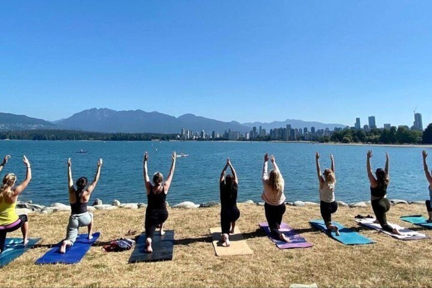 A group of people doing yoga on mats by a lake with a city skyline in the background. 2