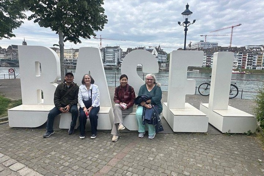 People sitting on benches in front of large white letters spelling 'BASEL'. 1