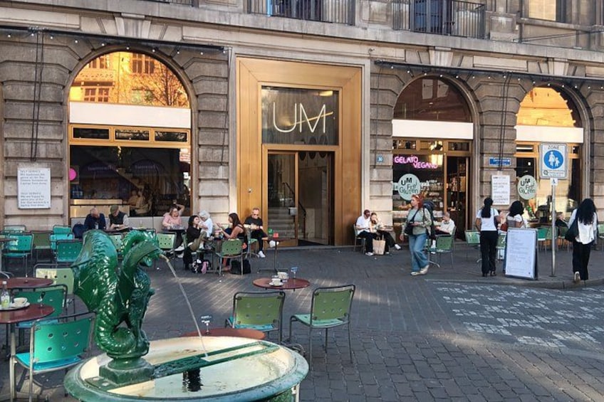 An outdoor café with tables and chairs, a decorative fountain, and storefronts with signage. 3