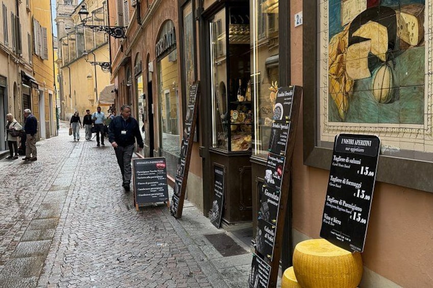 En kullerstensgata med folk som promenerar, butiker och en skylt med priser. 3