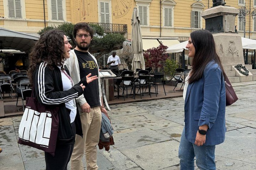Tre personer står utomhus, ett café med bord och parasoller syns, och i bakgrunden står en staty. 4